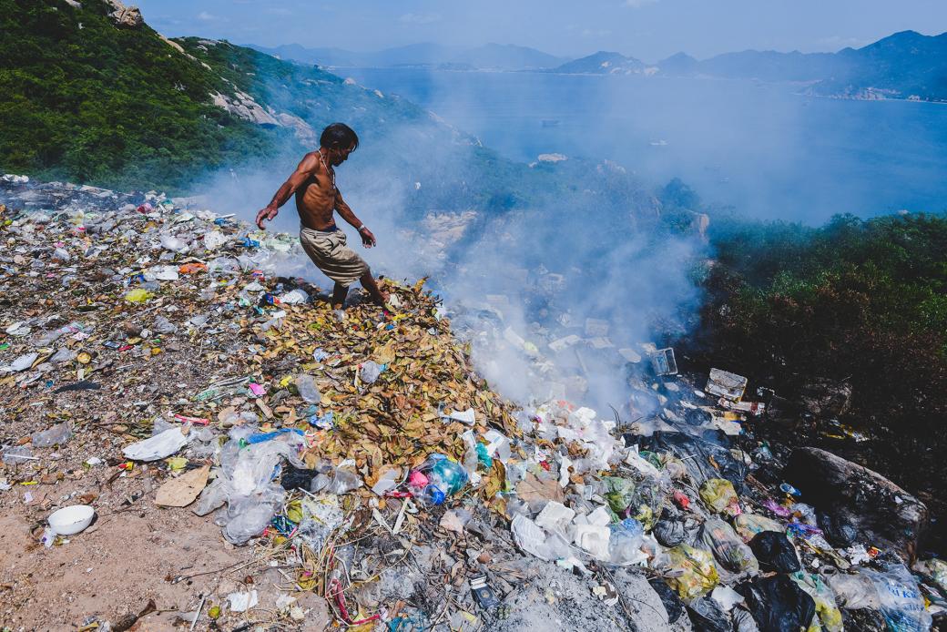 Ông Nguyễn Lương, người phụ trách lò đốt rác ở đảo Bình Ba (Khánh Hòa) đang dùng chân đẩy rác. Xung quay cấy cối chết khô. Đây là một sườn dốc của đảo, phía dưới là bãi tắm và là vịnh nuôi tôm hùm, thuỷ hải sản.