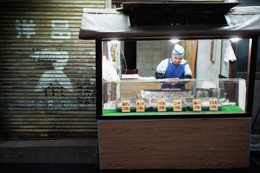 Cụ ông bên xe bán yakitori (gà xiên) truyền thống về đêm đứng trước một cửa hàng hiện đại tại Komagome.