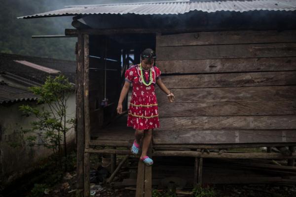 Một cô gái thiếu niên người bản địa đang bước ra khỏi ngôi nhà làm bằng gỗ lợp tole kẽm của gia đình, được xây dựng theo lối kiến trúc truyền thống.